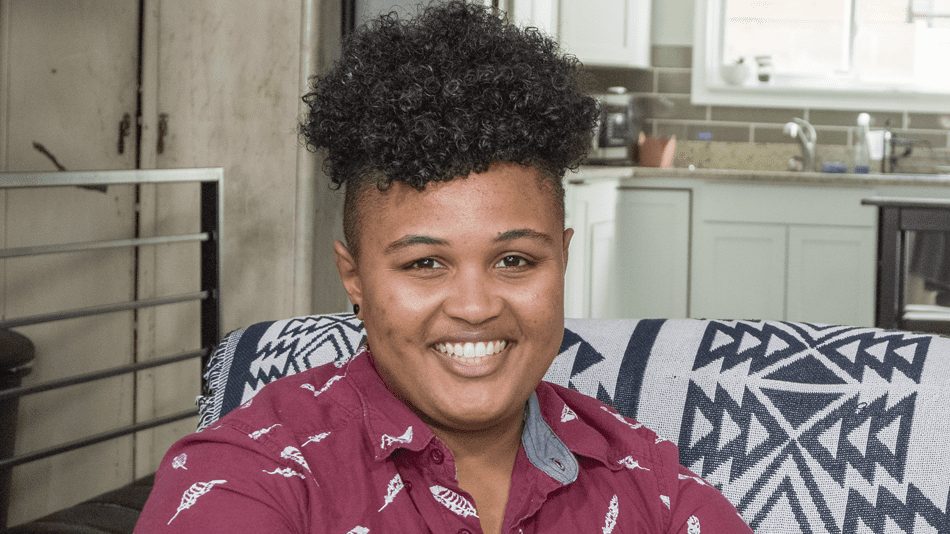 Grey, a Black queer person sits for interview on blue and white couch. They are smiling and wearing a red collared shirt with white embroidery in the shape of feathers