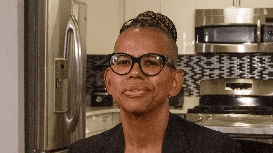 Headshot of Shellby, a Black trans nonbinary person. They are sitting in for their interview in a black blazer, wearing glasses and their braided hair is in a high bun.